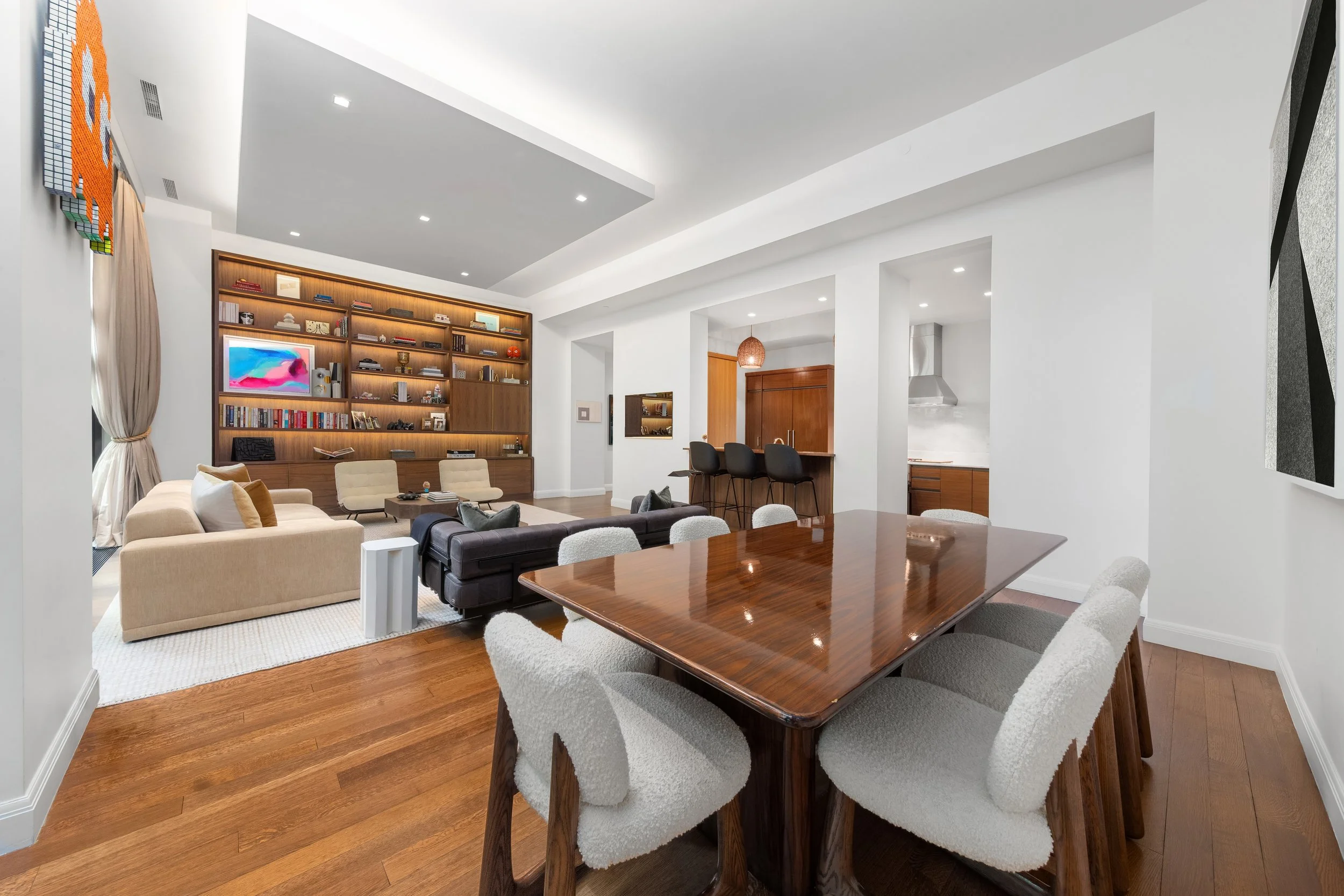 Modern open-plan living and dining room with white walls, wooden floors, a large wooden dining table with white chairs, a beige sofa, a dark brown sofa, a built-in wooden bookshelf, and a kitchen in the background.