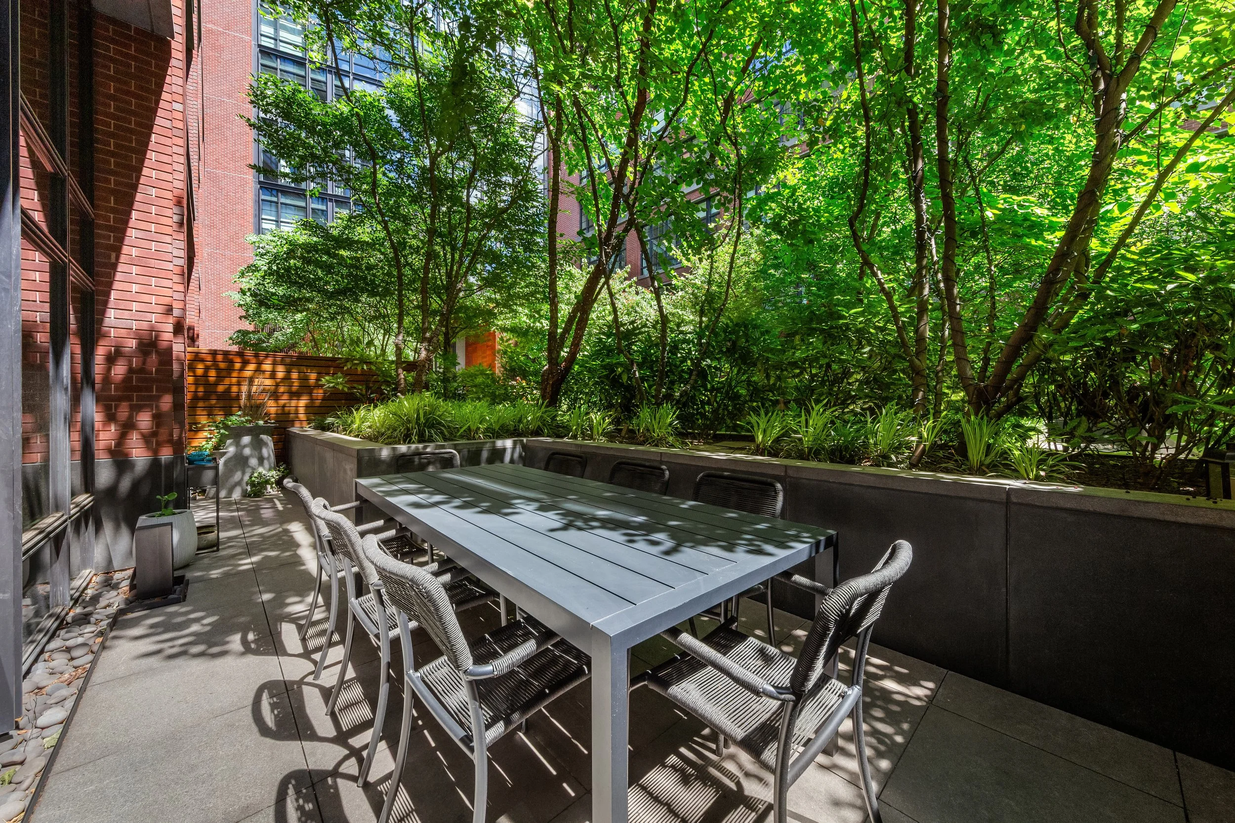Outdoor patio with a long table and wicker chairs, surrounded by lush green trees and plants, with building windows visible in the background.