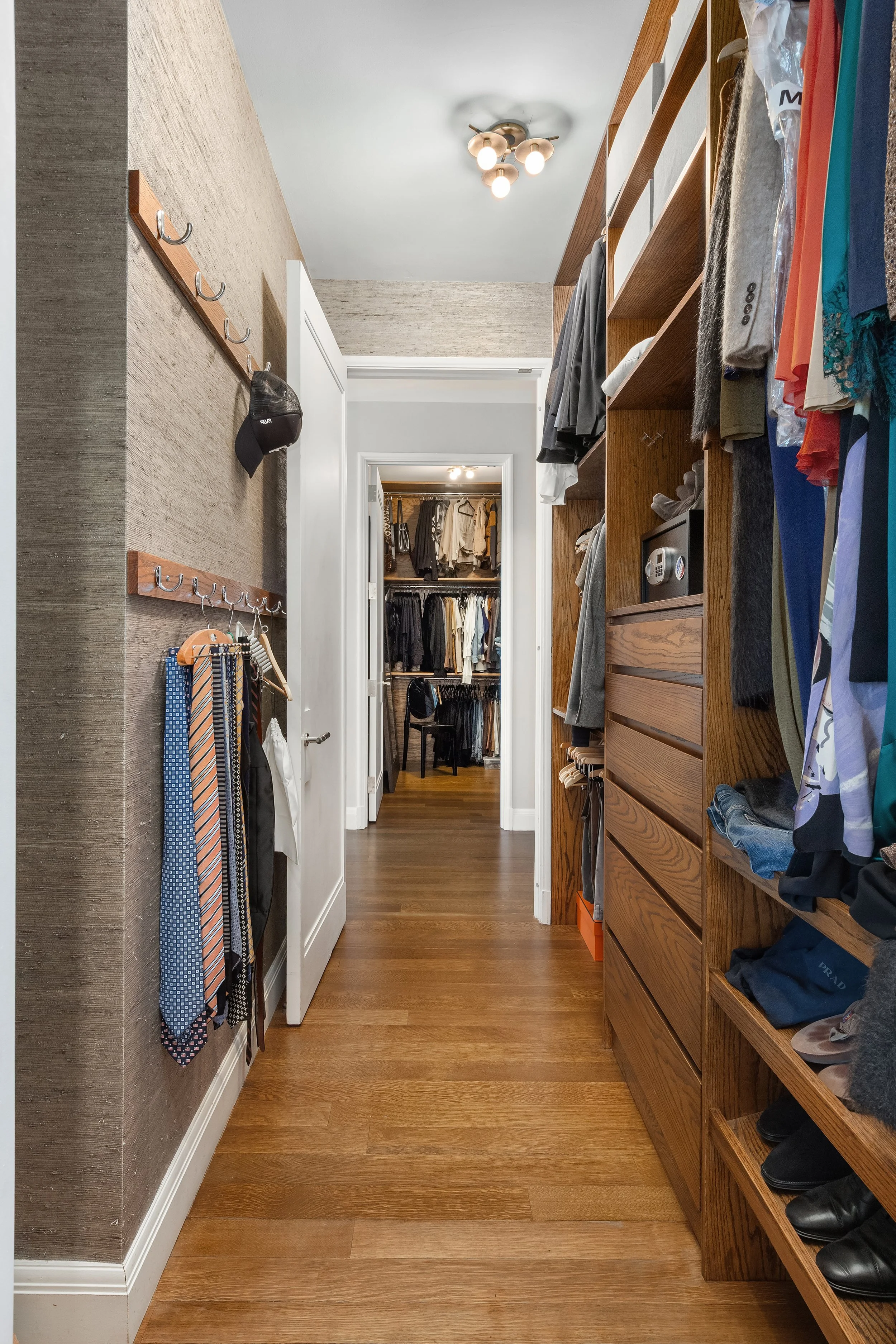 A walk-in closet with wooden shelves filled with folded clothes, shoes, and hanging garments. A doorway leads to another room with more clothing. The ceiling has a modern light fixture, and the floor is hardwood.