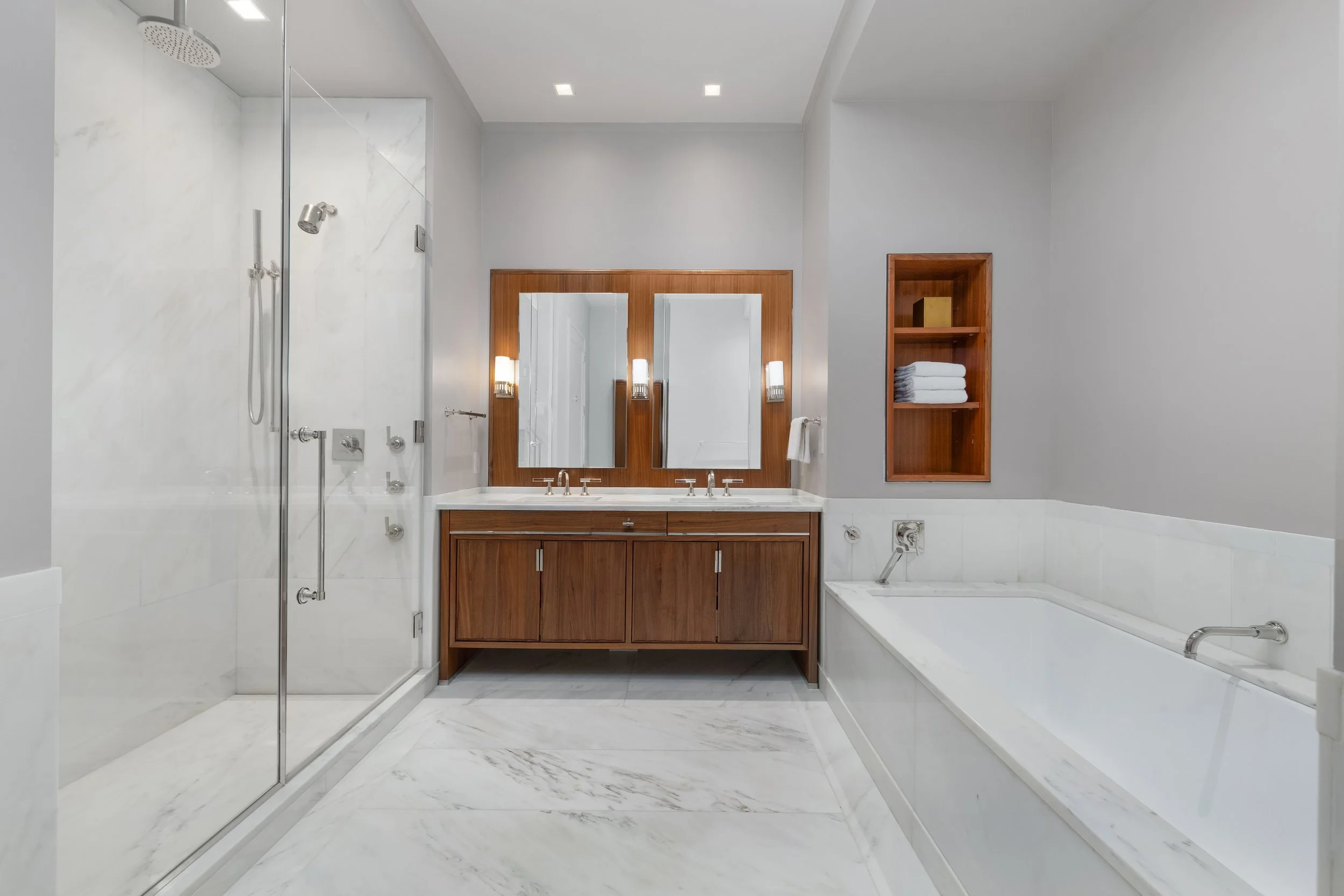 Modern bathroom with a glass-enclosed shower, a double sink vanity with wooden cabinets, large mirrors, a built-in bathtub, and shelves with towels.