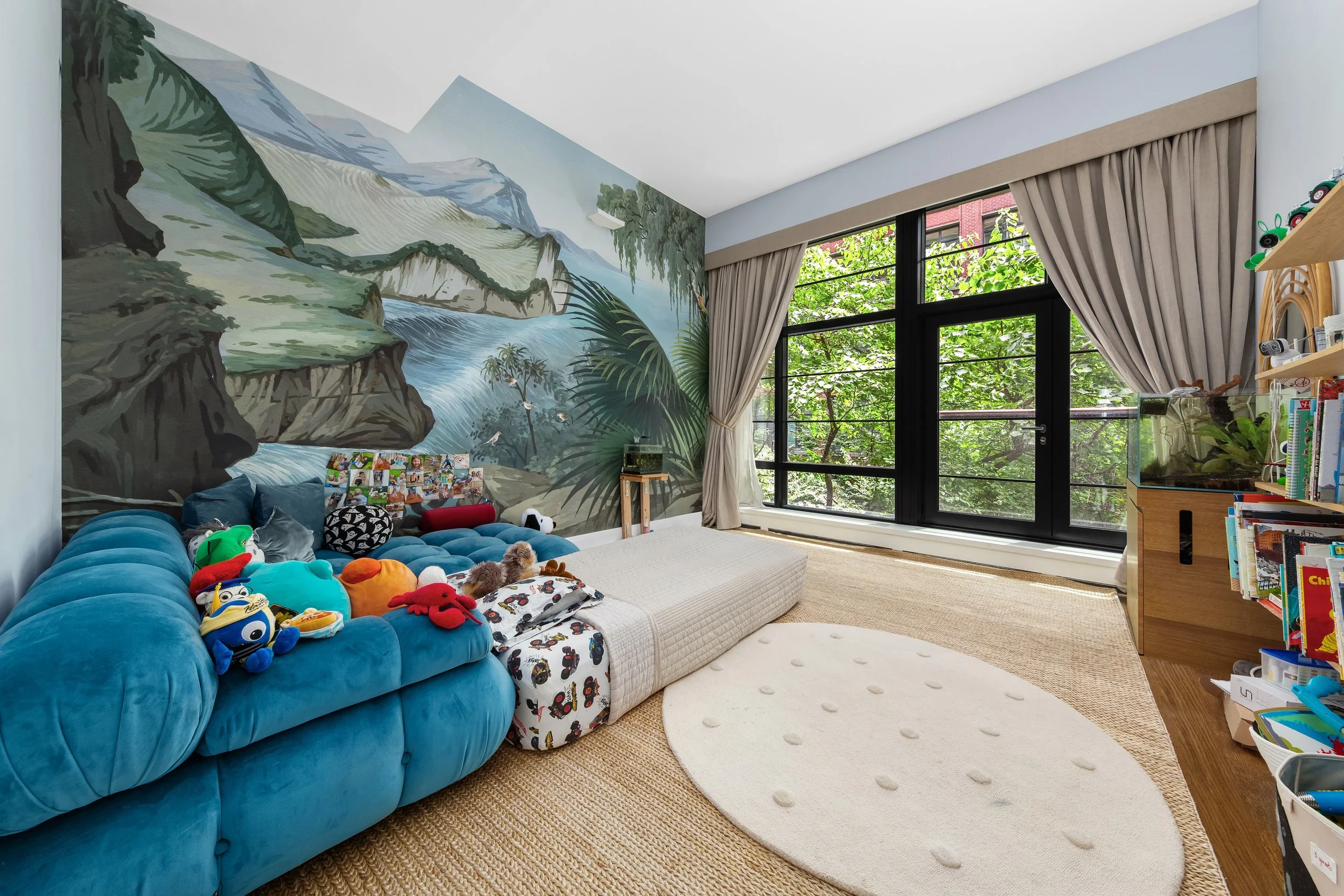 A brightly lit living room with a large blue sofa full of plush toys, a mural of mountains and rainforest on the wall, large windows with beige curtains, and a white rug on a woven carpet.