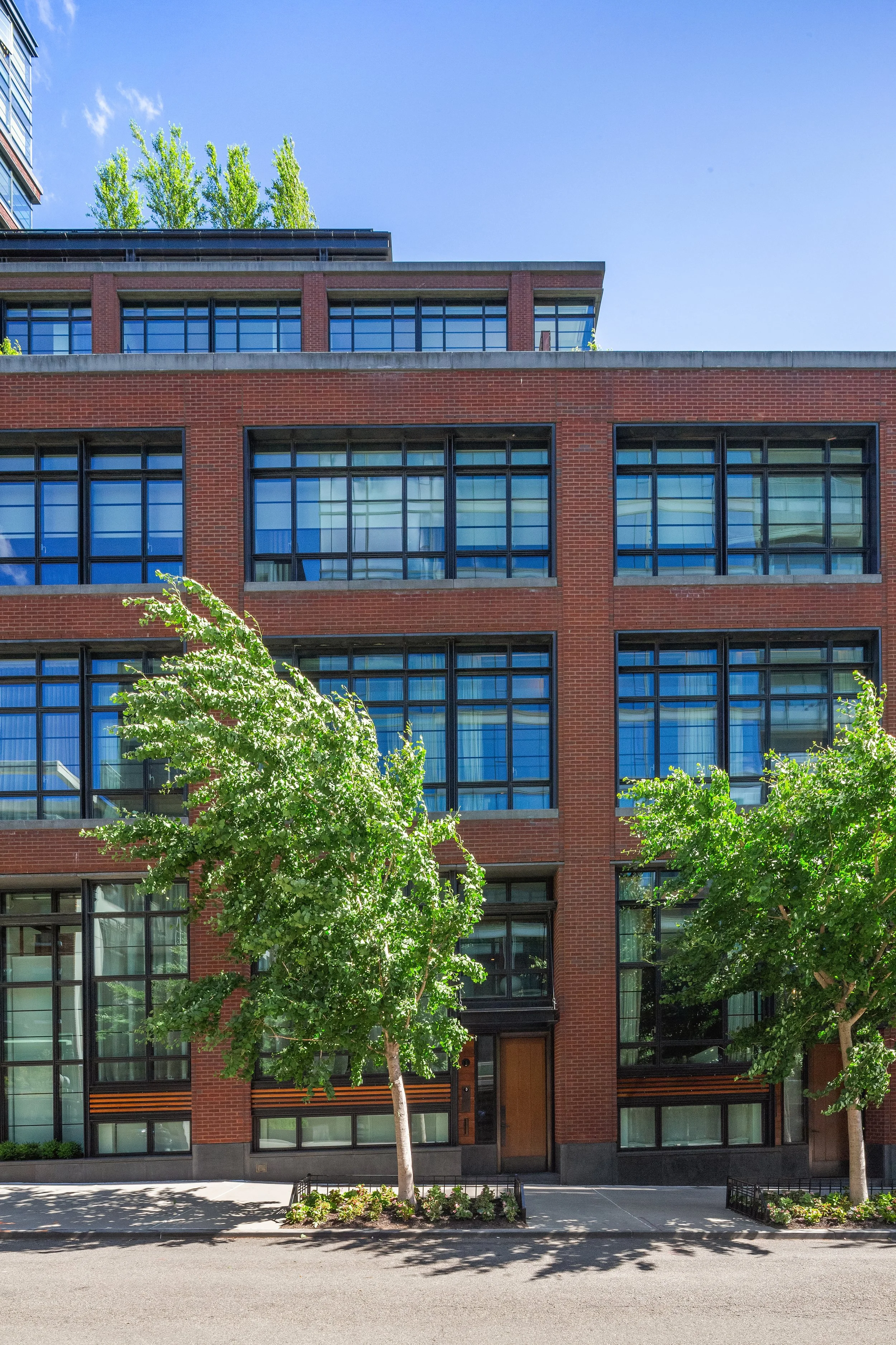 A modern multi-story red brick building with large rectangular glass windows and a flat roof. There are trees with green leaves in front of the building, a sidewalk, and a clear blue sky.