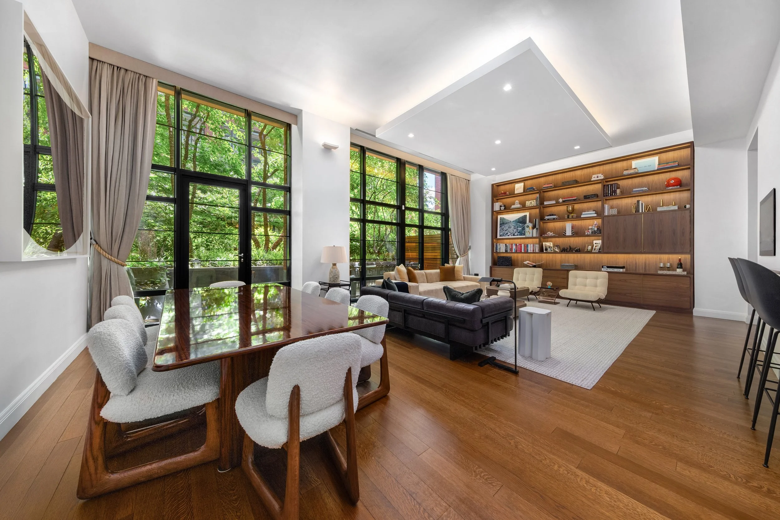 Modern living room with large windows, hardwood floors, white walls, and built-in wooden shelving. Contains a dining table with six white upholstered chairs, a beige sofa, black and beige armchairs, and a white side table. Decor includes books, frame