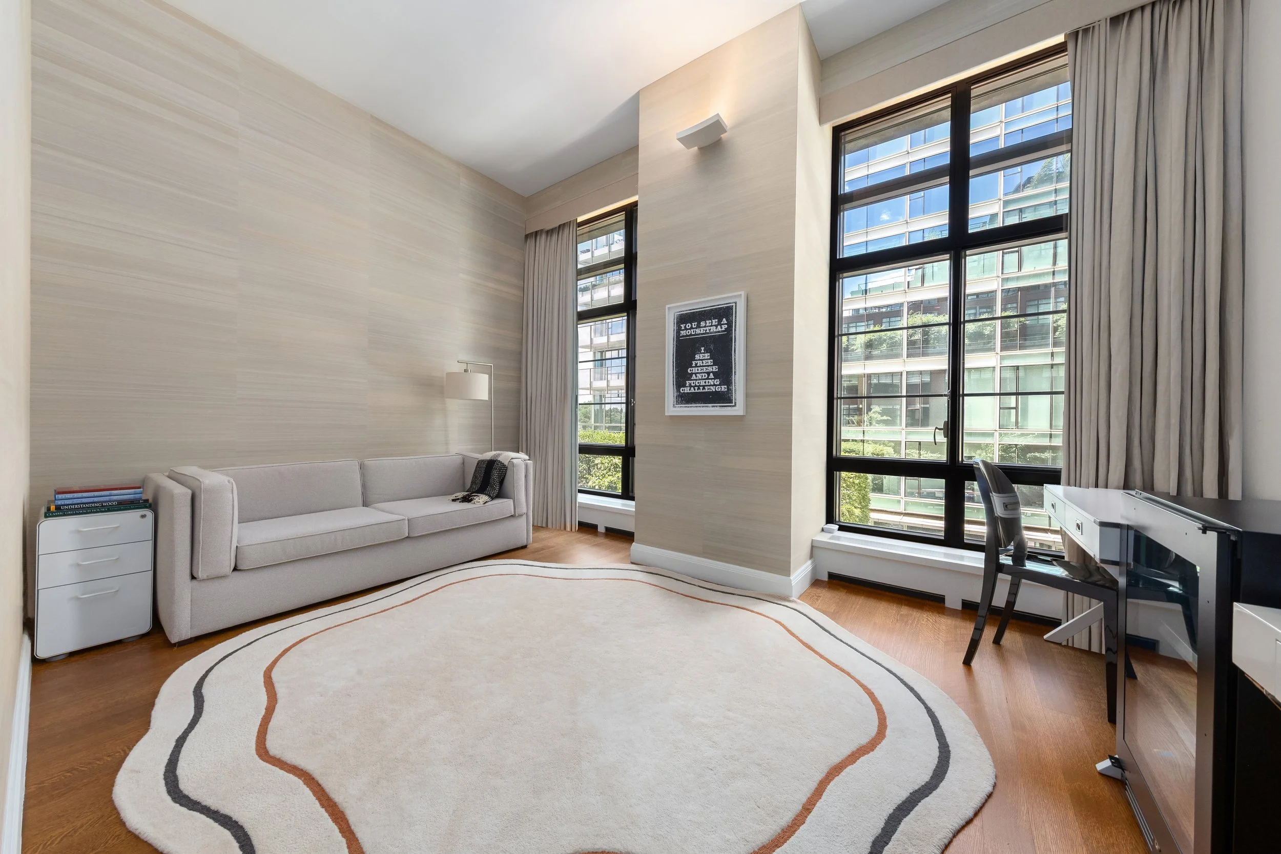 Modern living room with large windows, beige walls, a white sofa, a small white side table with books, a floor lamp, a patterned area rug, and a desk with an office chair.