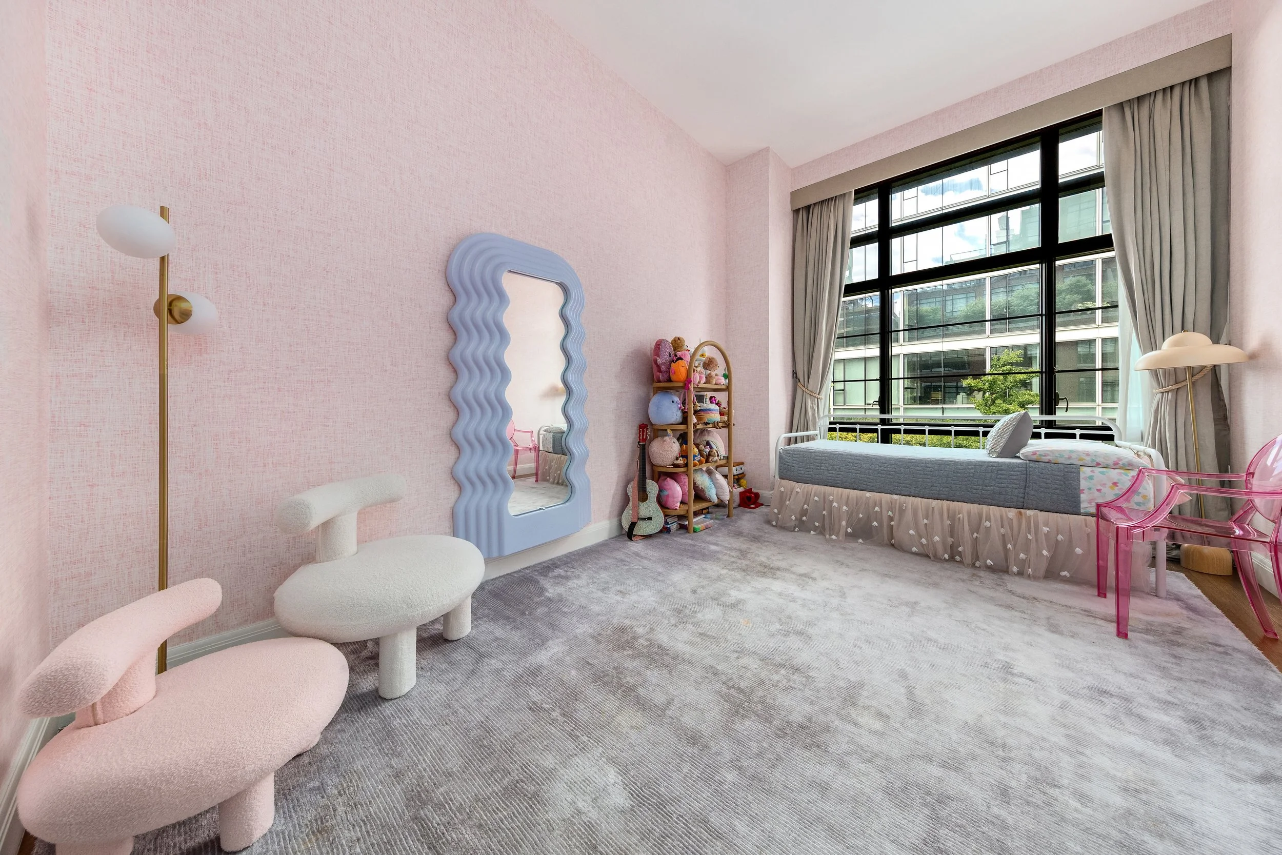 A cozy bedroom with pink walls, a large window with gray curtains, a white bed with stuffed animals, a pink transparent chair, a white chair with curved armrests, a tall gold floor lamp with white globes, a wavy-edged mirror with a blue frame, a wood