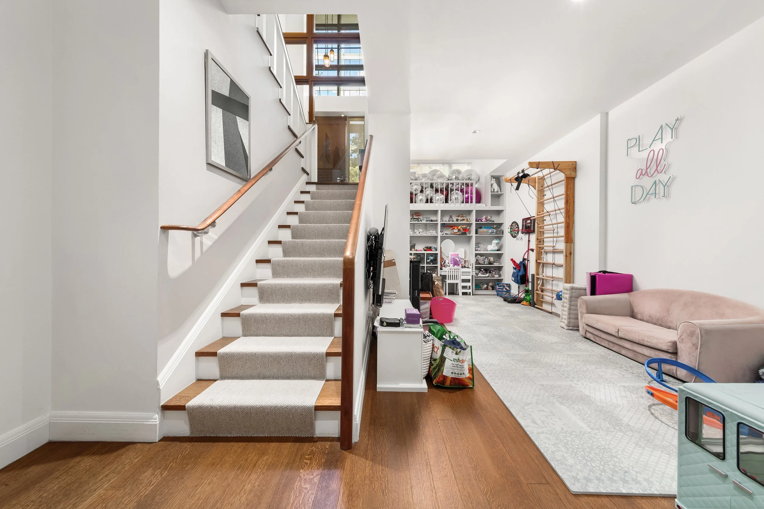 Interior of a modern living space with a staircase, a sitting area with a beige sofa, a wall sign saying 'PLAY all DAY,' and various toys and storage units.