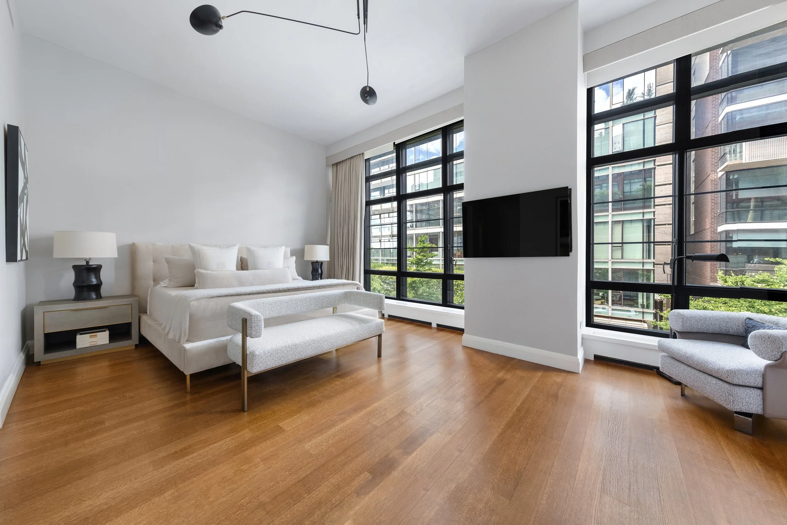 Modern living room with white walls, wooden floors, large windows with black frames, a white couch, black and white lamps, and a wall-mounted flat-screen TV.