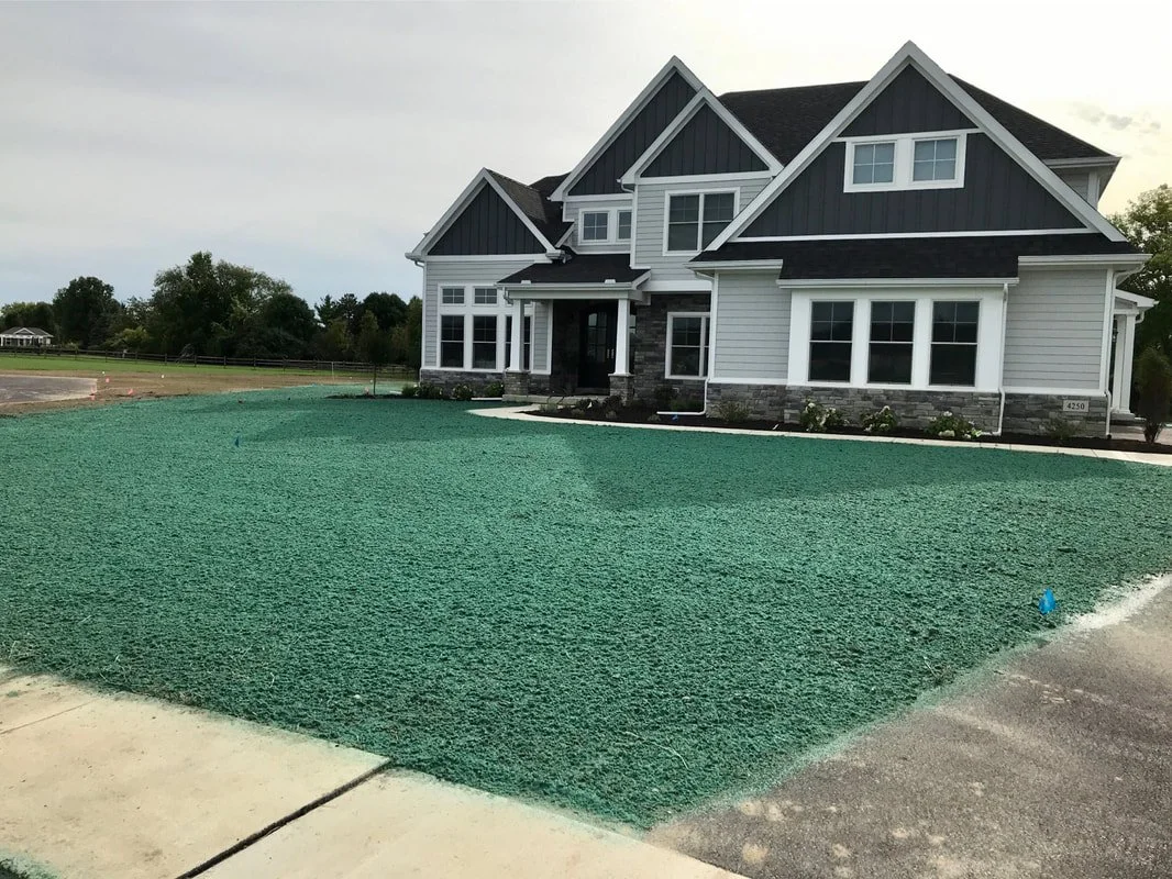 Newly constructed house with white siding, black roof, and large windows, situated on a lot with a newly laid green artificial turf yard.