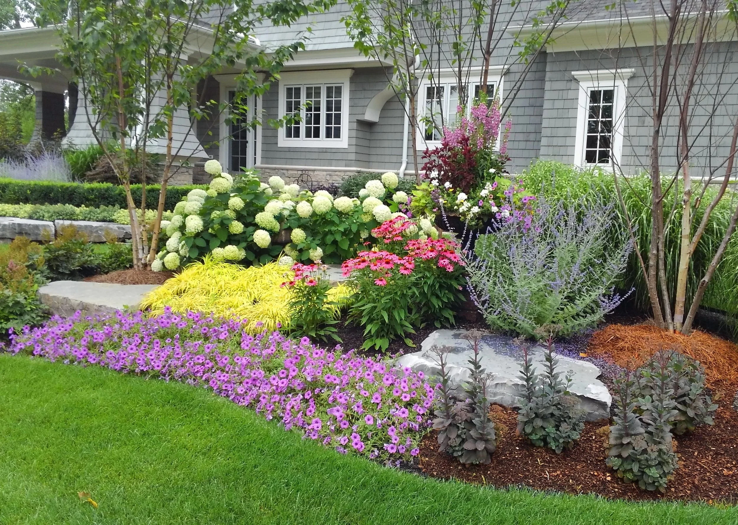 A front yard garden with various colorful flowers and green grass, in front of a gray house with white window frames and a stone foundation.