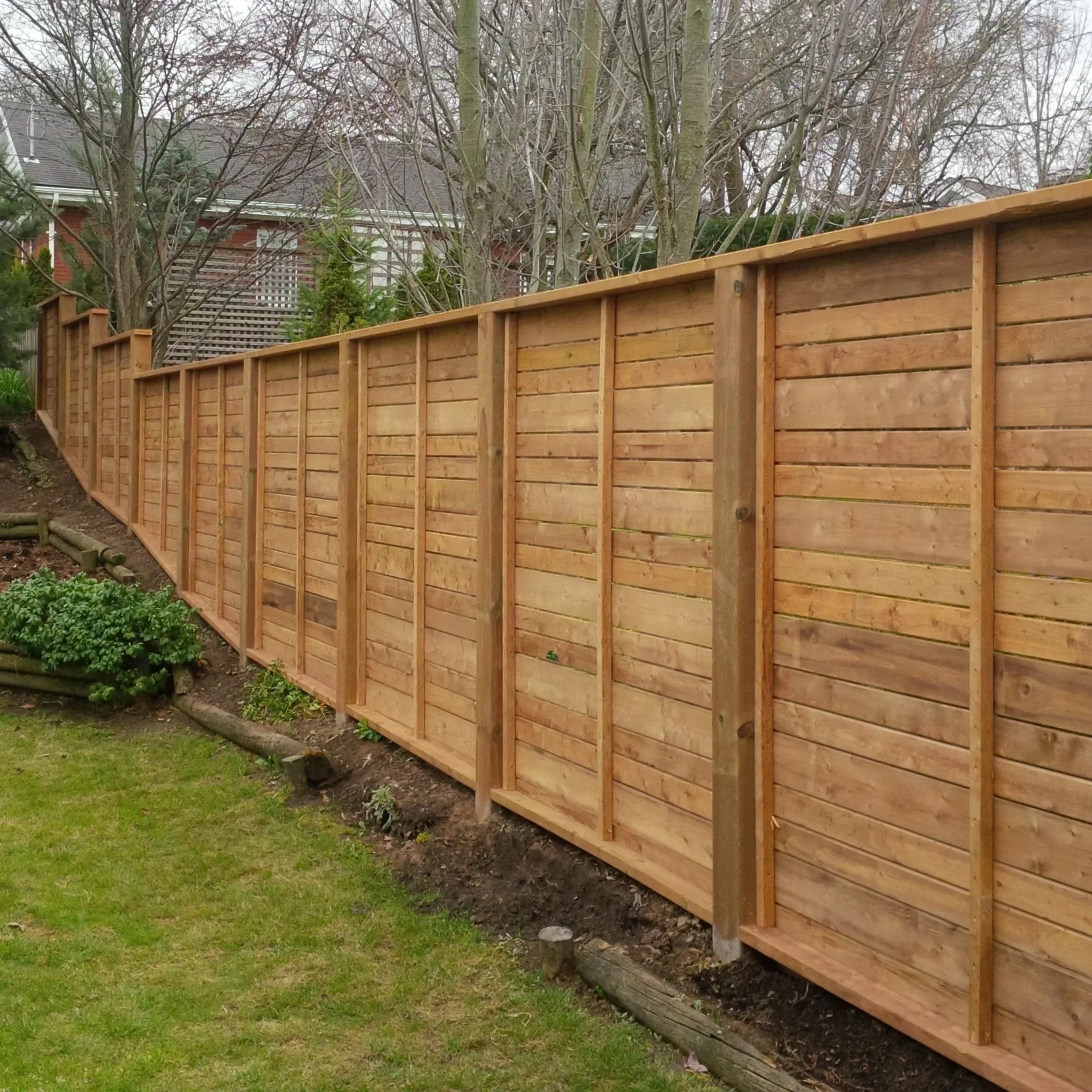A newly built wooden fence in a backyard with a hedge, leaving a small gap at the bottom near the soil. In the background, leafless trees and a house with a gabled roof are visible.