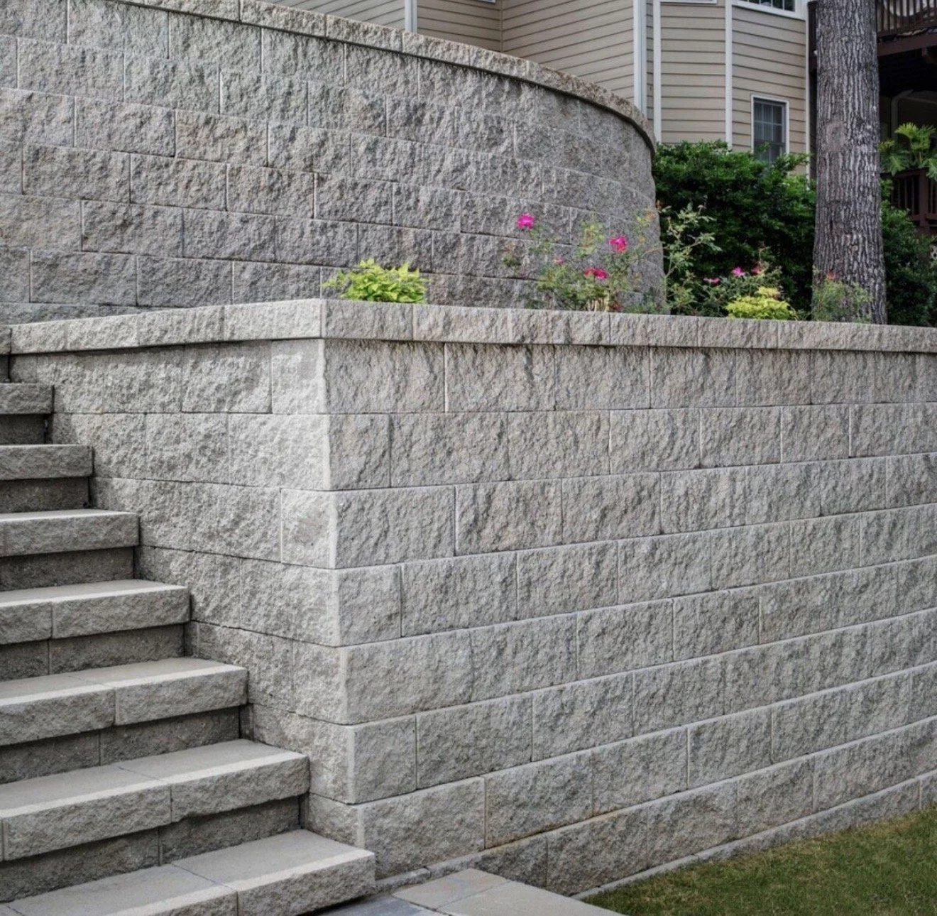 Stone retaining wall and stairs in a backyard with some pink and yellow flowers and a large tree.