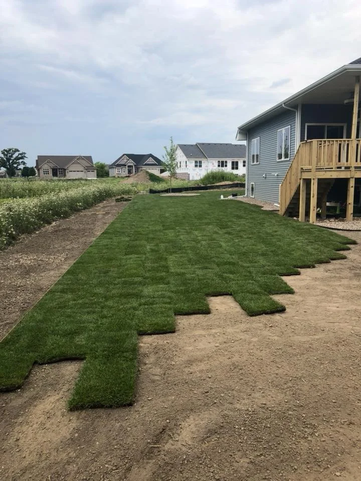 Artificial grass patches being installed on a lawn next to a gray house with a wooden deck, with other new houses visible in the background under a cloudy sky.