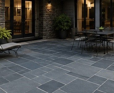 Outdoor patio with stone tile flooring, black metal furniture, potted plants, and illuminated windows of a building.