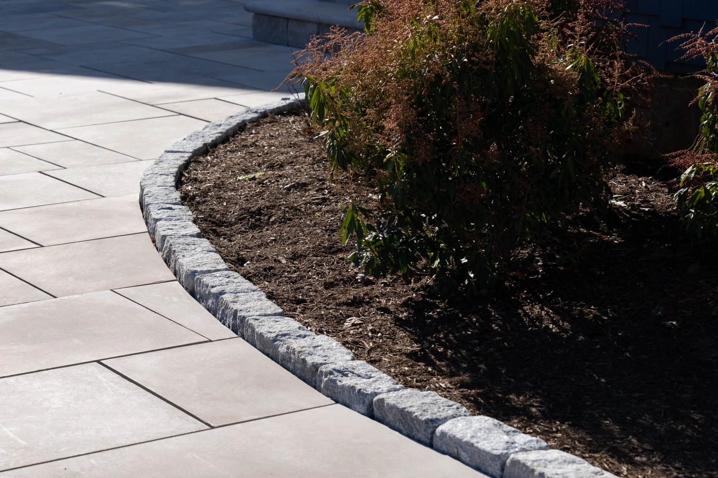 A sidewalk made of square beige tiles runs alongside a garden bed bordered by gray stones. The garden bed contains dark soil and some green leafy plants, with part of a dark-colored building in the background.
