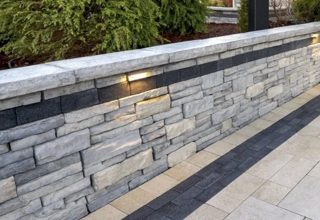 A stone decorative wall with built-in lighting, adjacent to a tiled walkway, with greenery in the background.