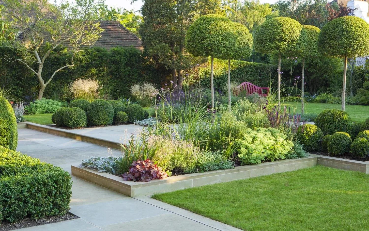 A beautifully landscaped garden with trimmed bushes, trees, and colorful plants, featuring a red bench and a walkway.