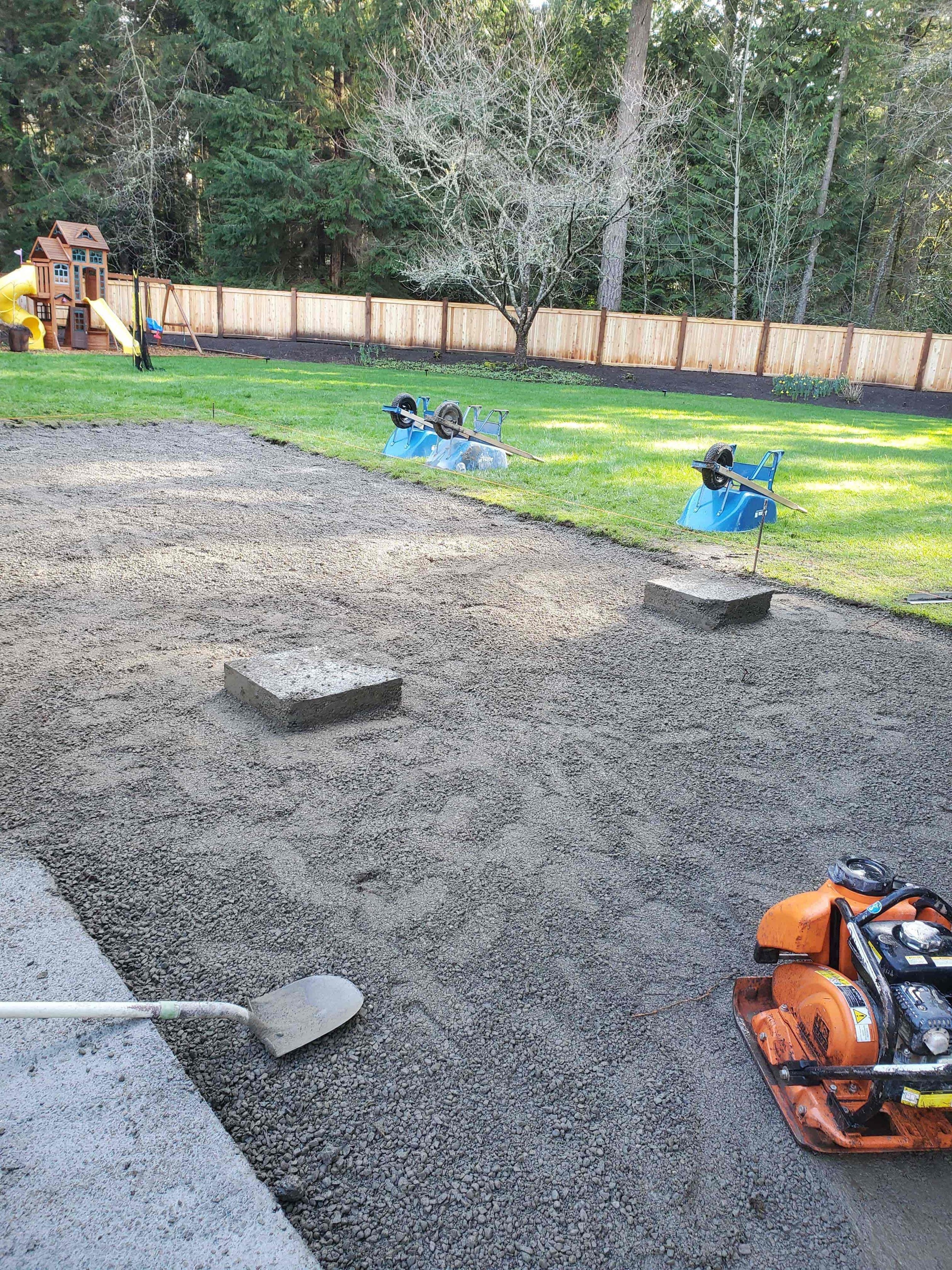 A backyard with a wooden fence, a playset with slides, a tree with no leaves, and a partially laid gravel pathway with construction tools including a shovel and a power tool.