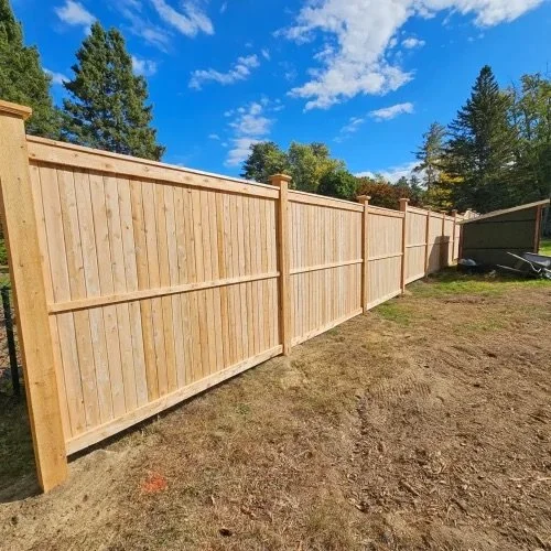 A newly built wooden fence in an open backyard with a clear blue sky and some trees in the background.