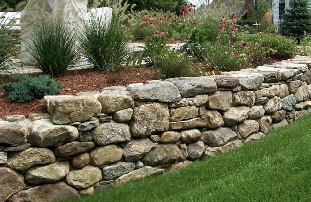 Stone retaining wall with plants and flowers on top, next to a green lawn.