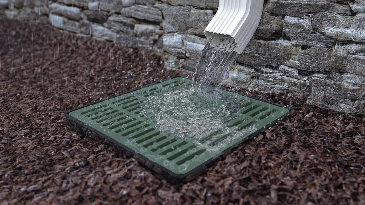 Water flowing from a downspout into a drain grate on a gravel surface next to a stone wall.