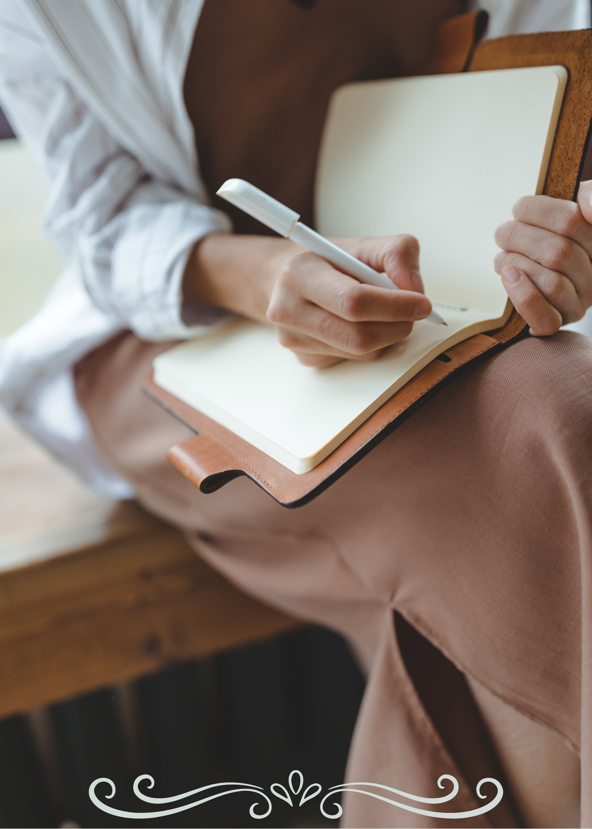 Close-up of a person wearing a white shirt writing in a cream-colored notebook with a white pen, seated at a wooden desk.