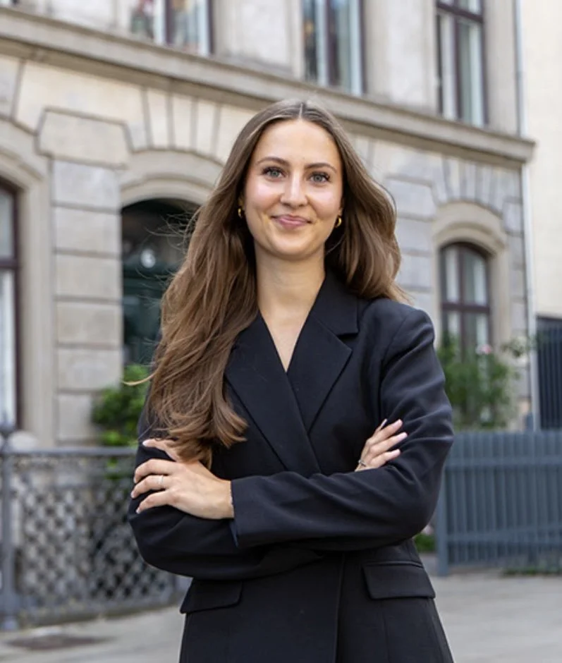 En ung kvinde med langt brunt hår, iført sort blazer, står foran en bygning med store vinduer og brolagte omgivelser.
