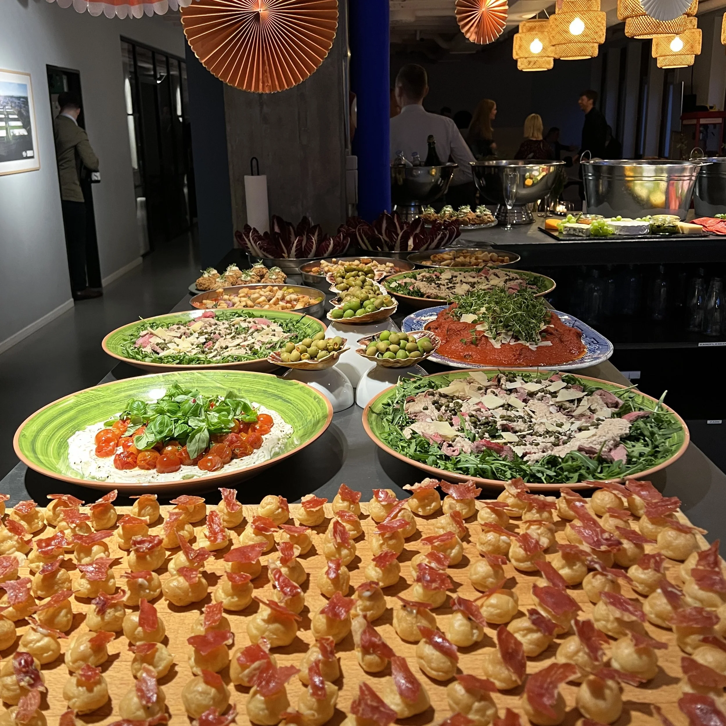 Et langt bord fyldt med små appetitvækkere. Bagved ses gæster, der snakker, og dekorative lampehoveder i varmt gult lys.