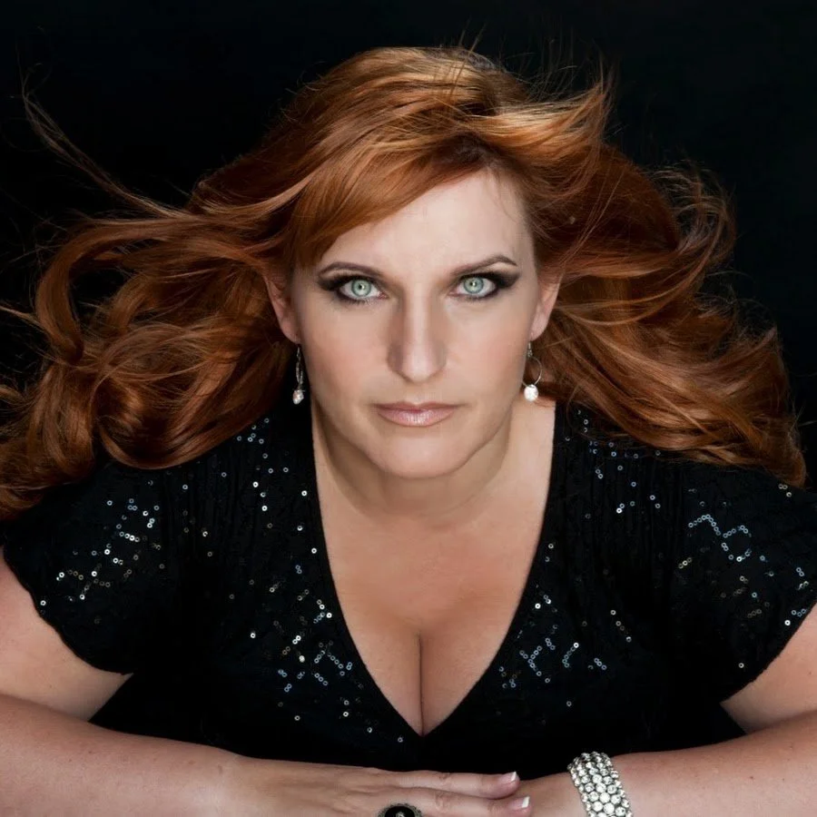 A woman with long red hair and blue eyes wearing a black sequin dress, pearl earrings, and a bracelet, lying down with her head facing the camera against a black background.