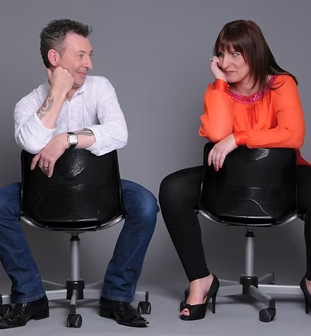 A man and a woman sitting on black office chairs facing each other on a gray background, with the man smiling and the woman looking at him with a playful expression.