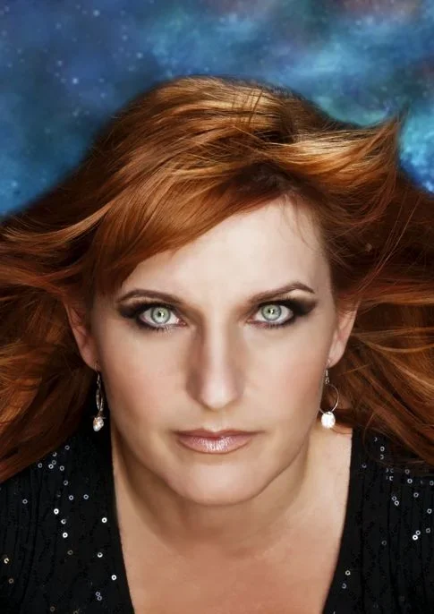 TJ Higgs with long red hair and piercing blue eyes, wearing earrings and a black, sequined top, against a cosmic blue and starry background.