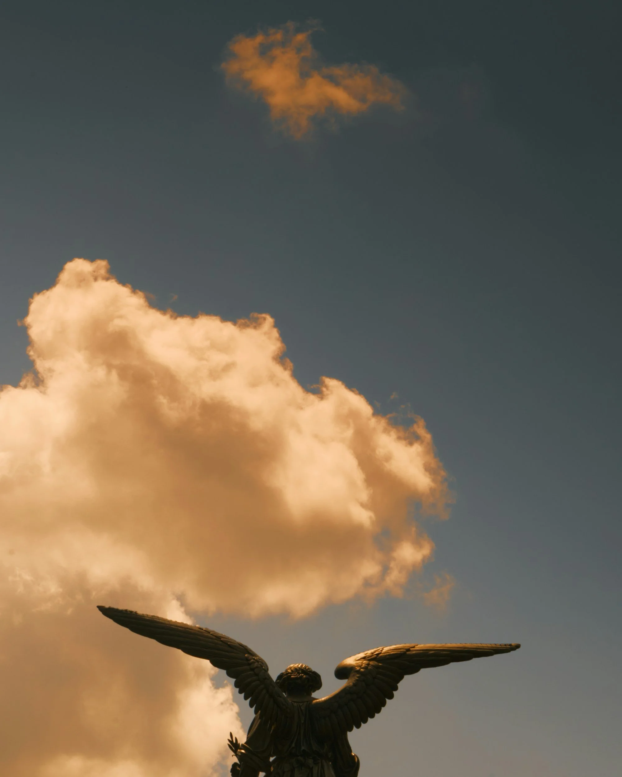 A bronze angel statue with outstretched wings stands against a cloudy sky during sunset, with the sun illuminating the clouds and the statue.