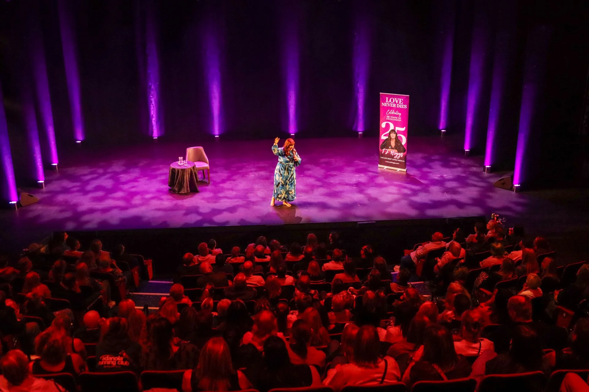 A woman with red hair on stage giving a speech or performance at an event celebrating 25 years of Mediumship, with an audience seated in front. The stage has purple lighting and a large pink vertical banner with text and a photo of the woman.
