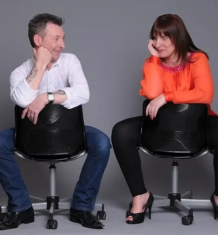 A man and woman sit on black chairs facing each other, smiling and making eye contact, set against a plain gray background.