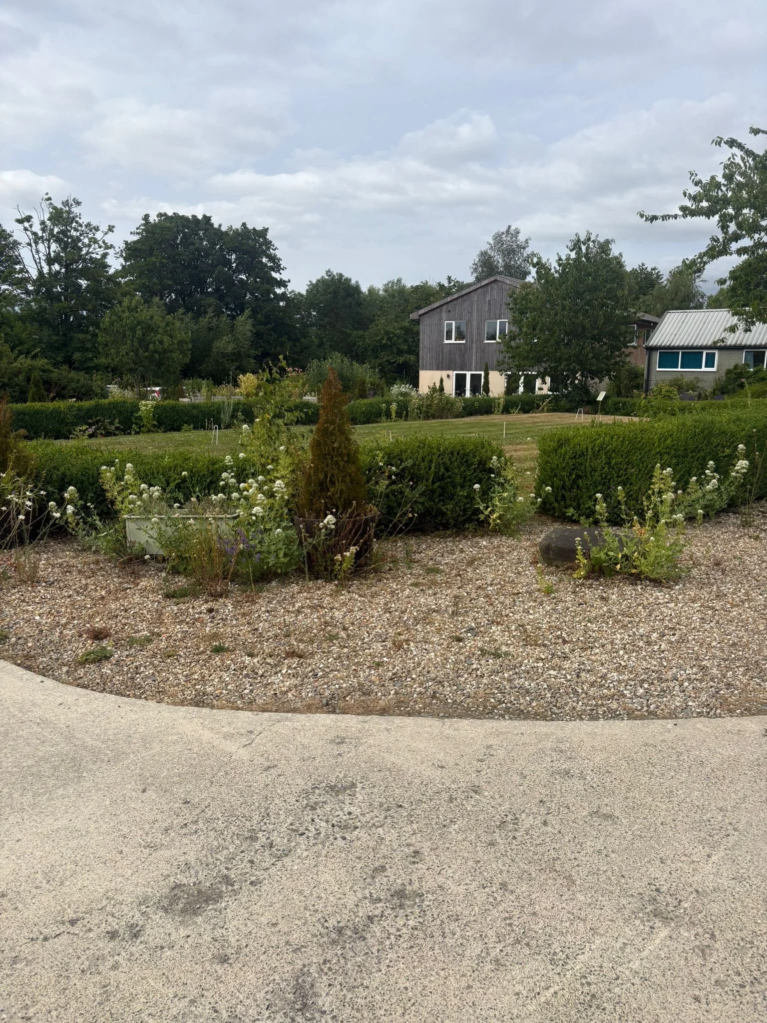 View of a yard with a gravel area in the foreground, maintained lawn, lush green bushes, and trees, with two modern houses in the background under a partly cloudy sky.