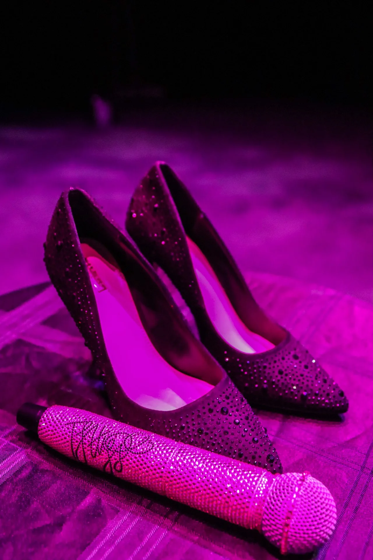 A pair of purple high-heeled shoes with rhinestone embellishments and pointed toes, placed on a surface under purple lighting. A decorated purple pen is in the foreground.