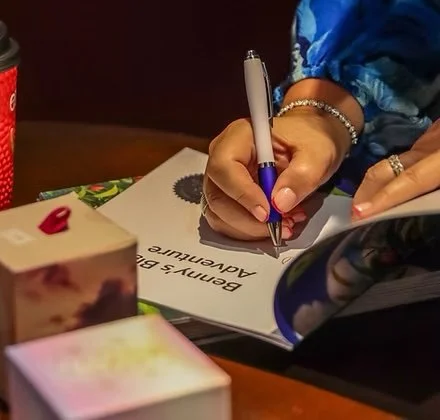 Person writing on a piece of paper with a pen, surrounded by books, a tablet, and a red coffee cup on a wooden table.