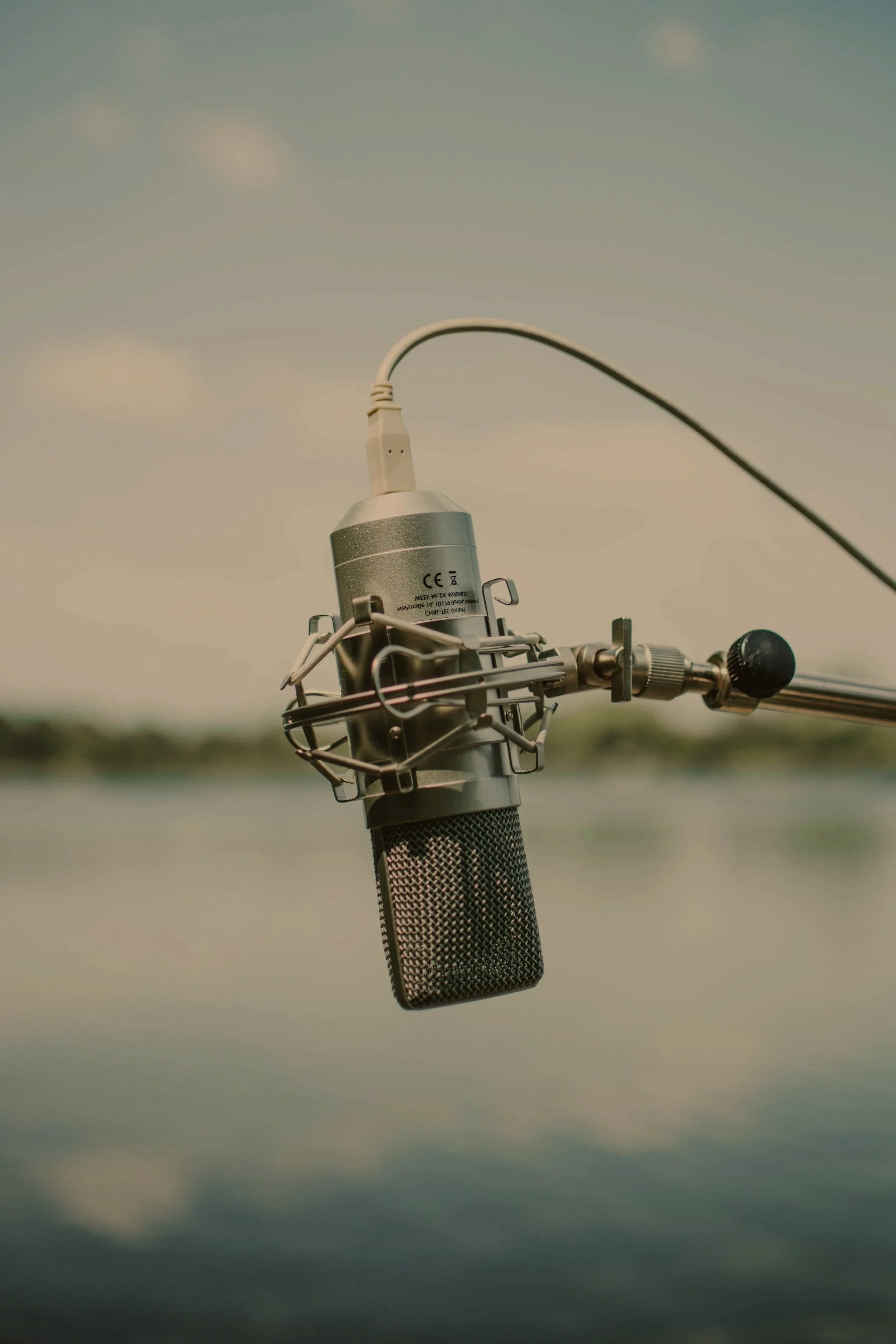 A vintage microphone with a mesh grille and metal frame, attached to a stand, against a cloudy sky background.