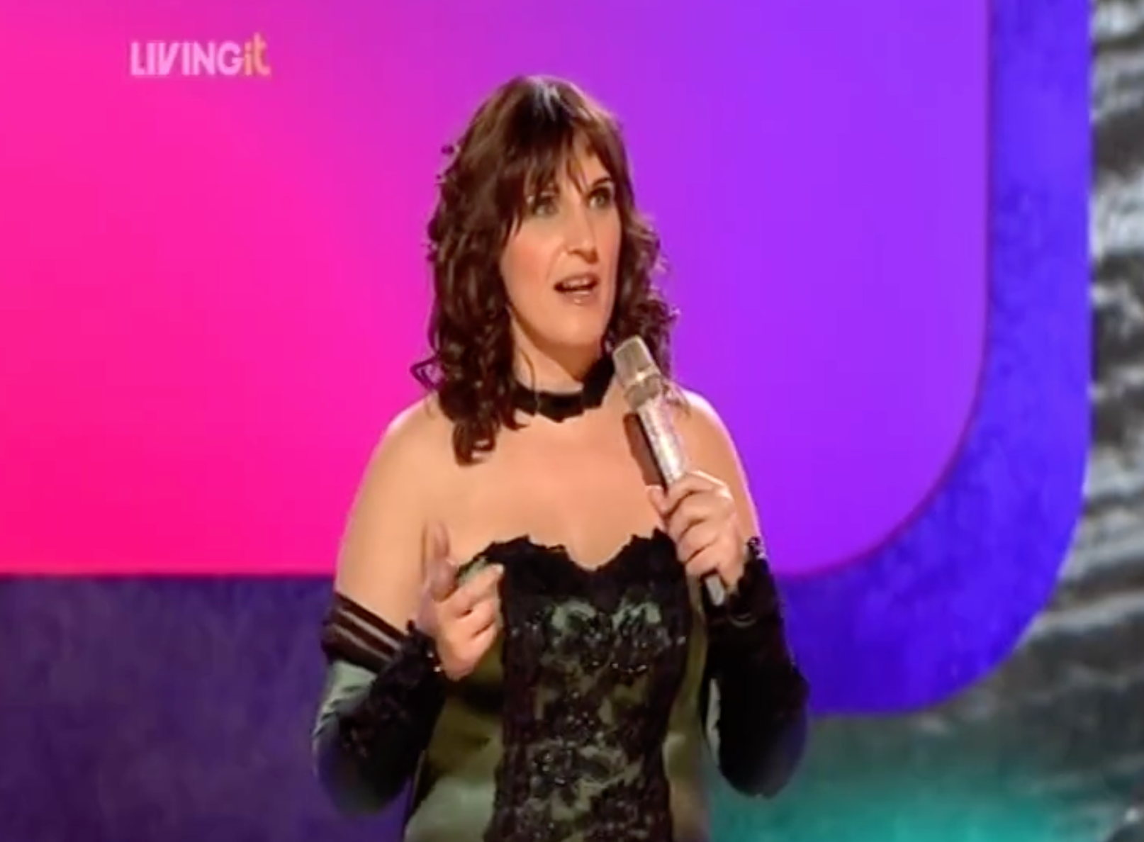 A woman with curly brown hair wearing a black lace off-shoulder dress and long black gloves, holding a microphone and speaking on a stage with a purple and pink background that has the text 'LIVING it'.