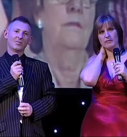 A man and a woman standing on stage holding microphones, with a large screen behind them displaying a close-up of a person's face.
