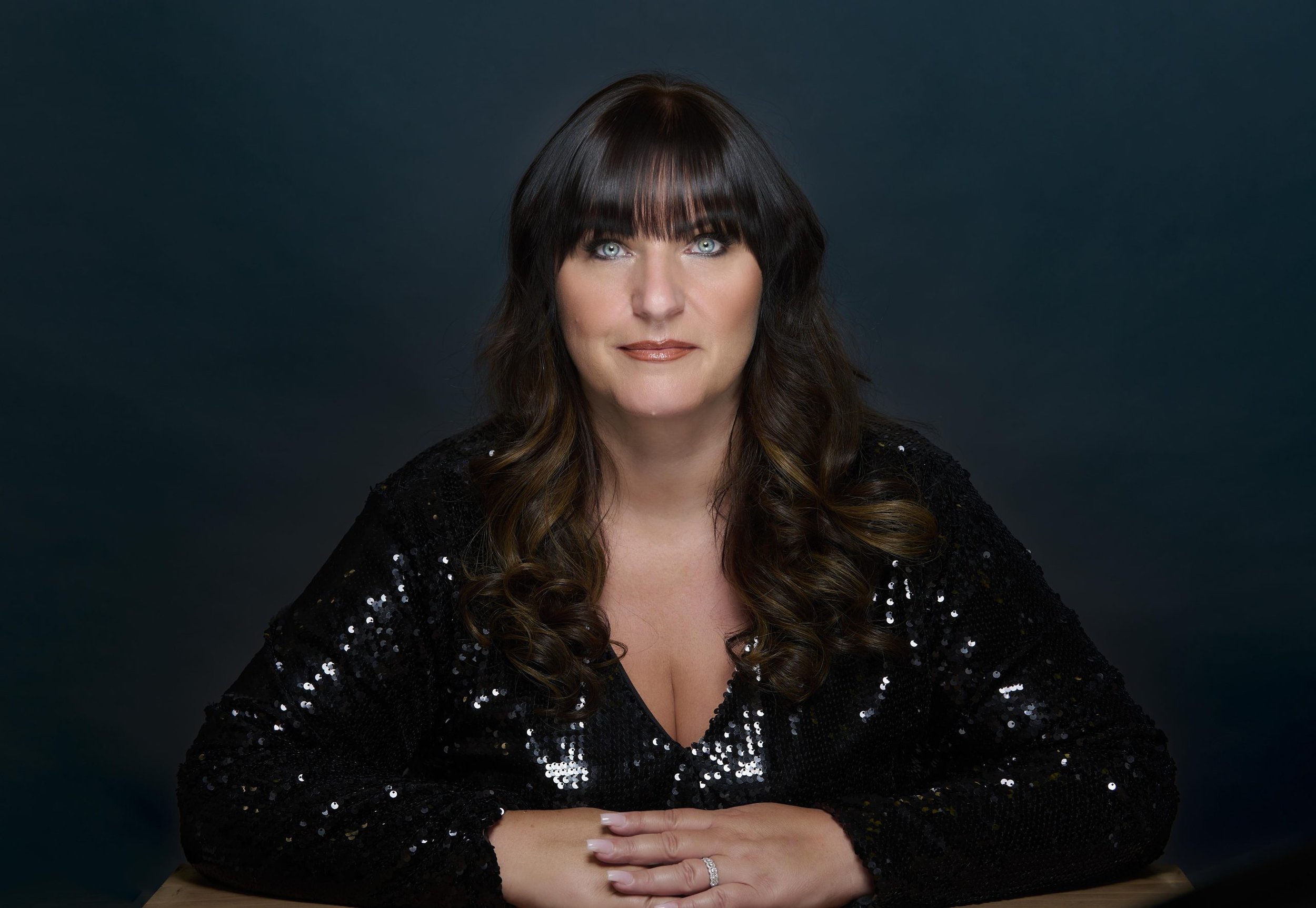 A woman with black hair and bangs, wearing a black sequin dress, seated with her hands folded on a wooden surface, against a dark background.