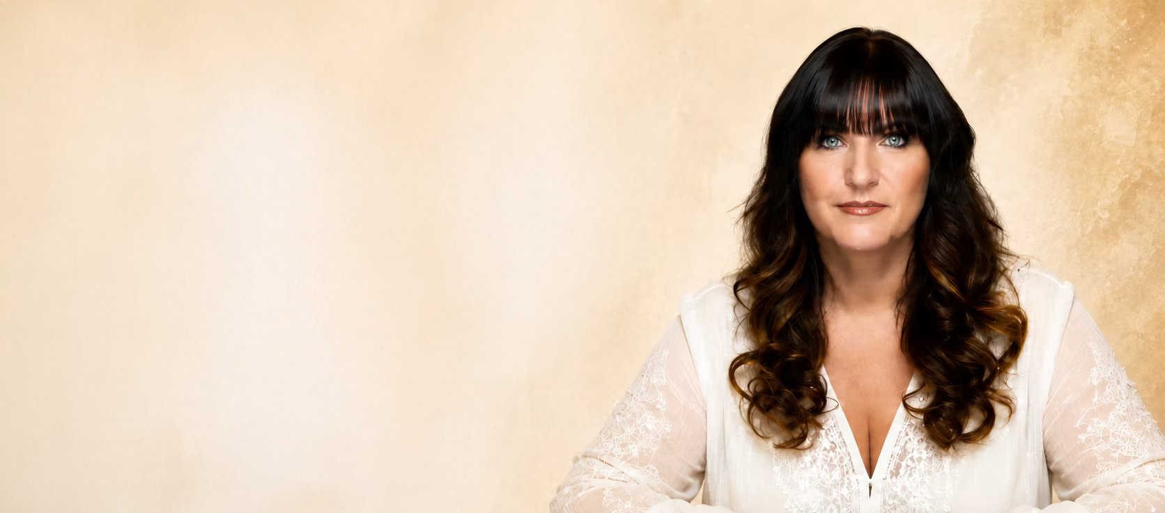 A woman with long dark curly hair and bright blue eyes, wearing a white top with lace sleeves, sitting against a light beige background.