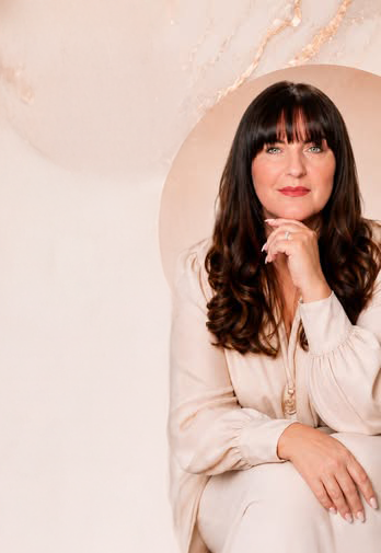A woman with long dark hair and straight bangs, wearing a light-colored outfit, sitting and smiling with her chin resting on her hand. She is positioned in front of a light-colored and marble-textured background.