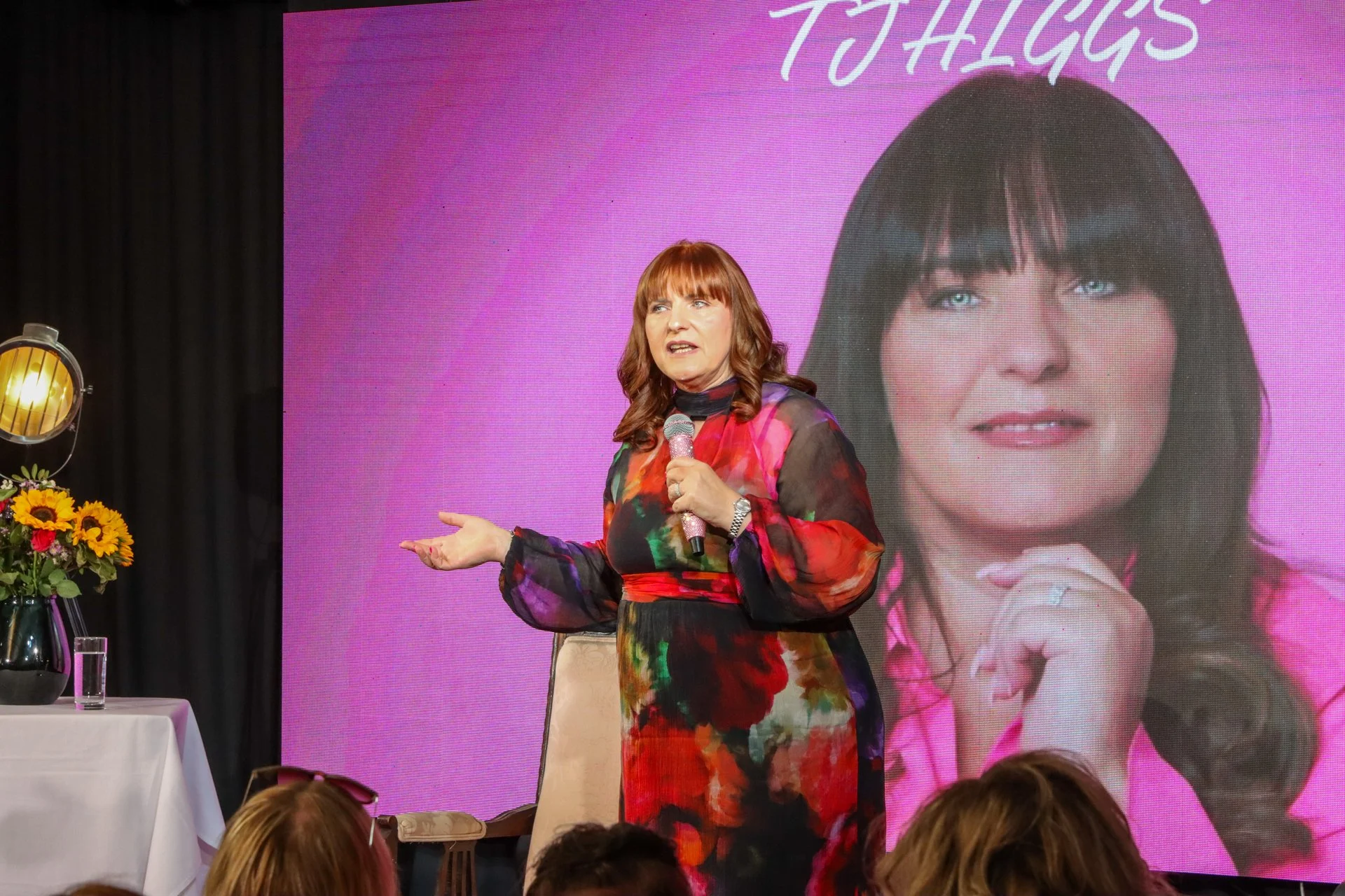 A woman with red hair and a colorful dress speaking into a microphone on stage, with a large pink screen behind her displaying her picture and the name 'TALIA GOSS'.