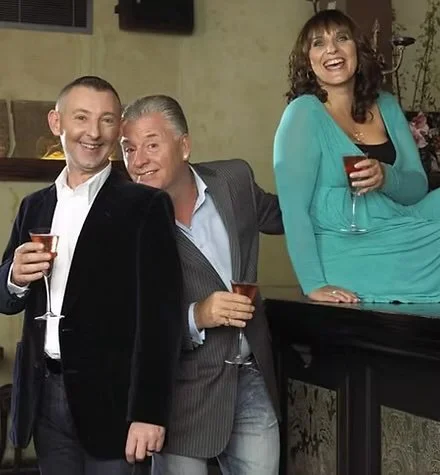 Three adults, two men and one woman, at a social gathering, holding glasses of wine, standing and leaning on a bar or counter, smiling and enjoying themselves.