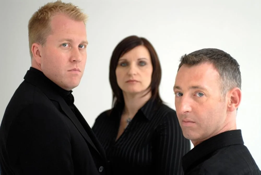 Three people, two men and one woman, standing against a plain white background, looking serious and intense.