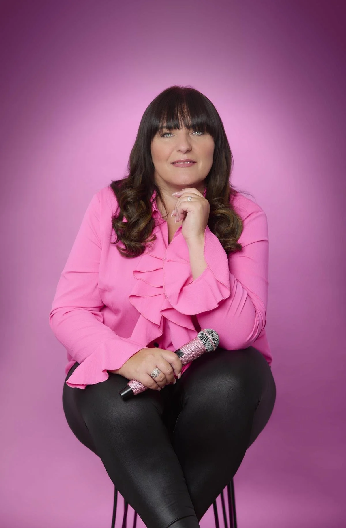 TJ with dark brown hair and bangs, wearing a pink ruffled blouse and black pants, sitting on a black stool against a pink gradient background. She holds a sparkly pink microphone in her right hand, with her left hand under her chin, smiling slightly.