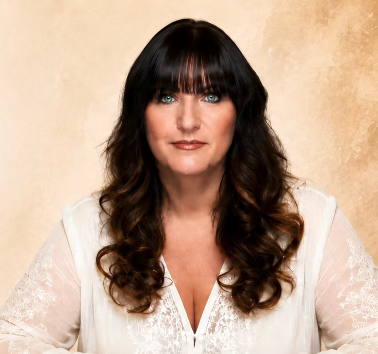 TJ Higgs with long brown hair and bangs, wearing a white blouse with lace details, looking directly at the camera against a neutral beige background.
