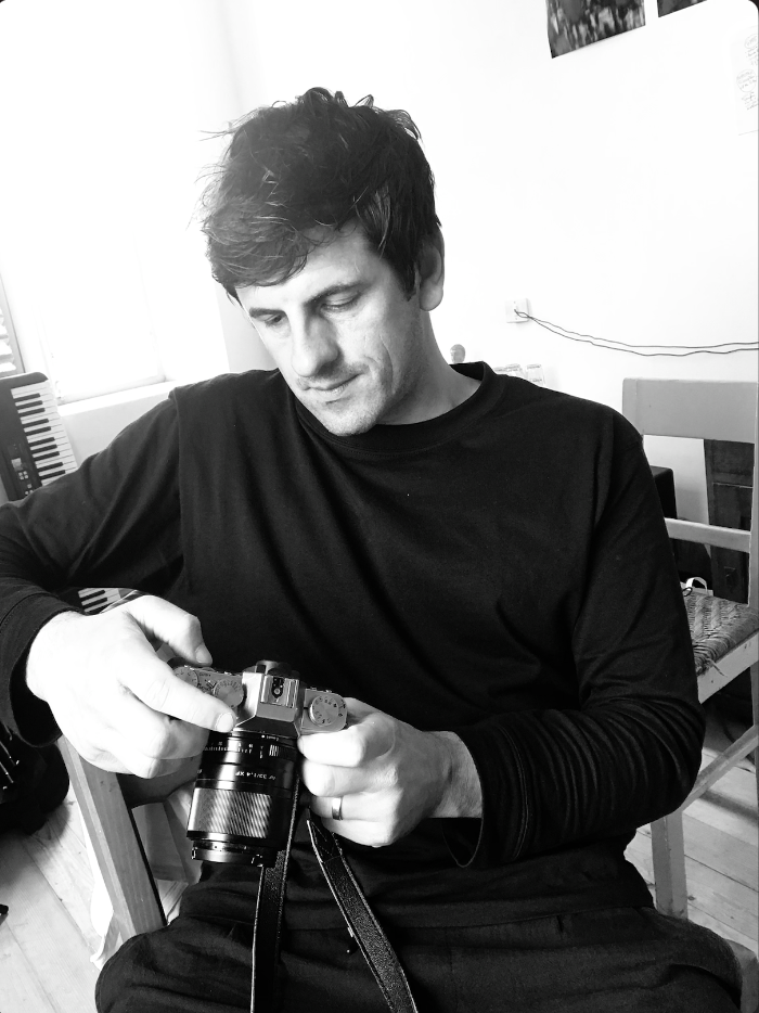 A man wearing a black shirt sitting in a room, looking at a camera he is holding.