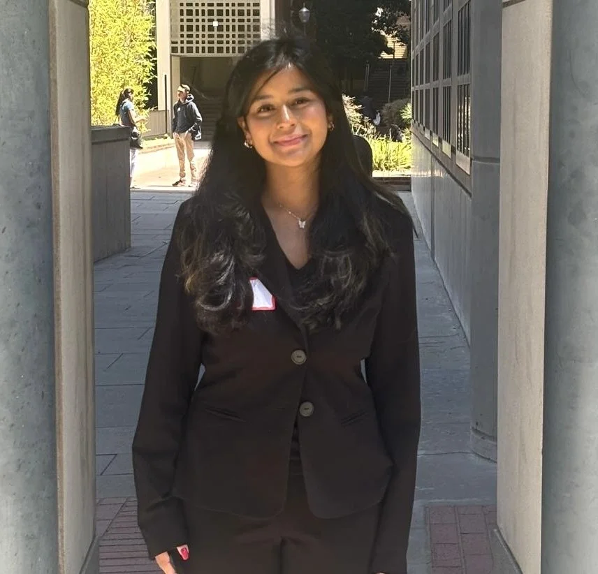 Young woman in black business suit standing outdoors, smiling, with people and trees in the background.