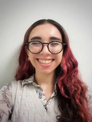 A young woman with glasses and long, wavy hair smiling at the camera.